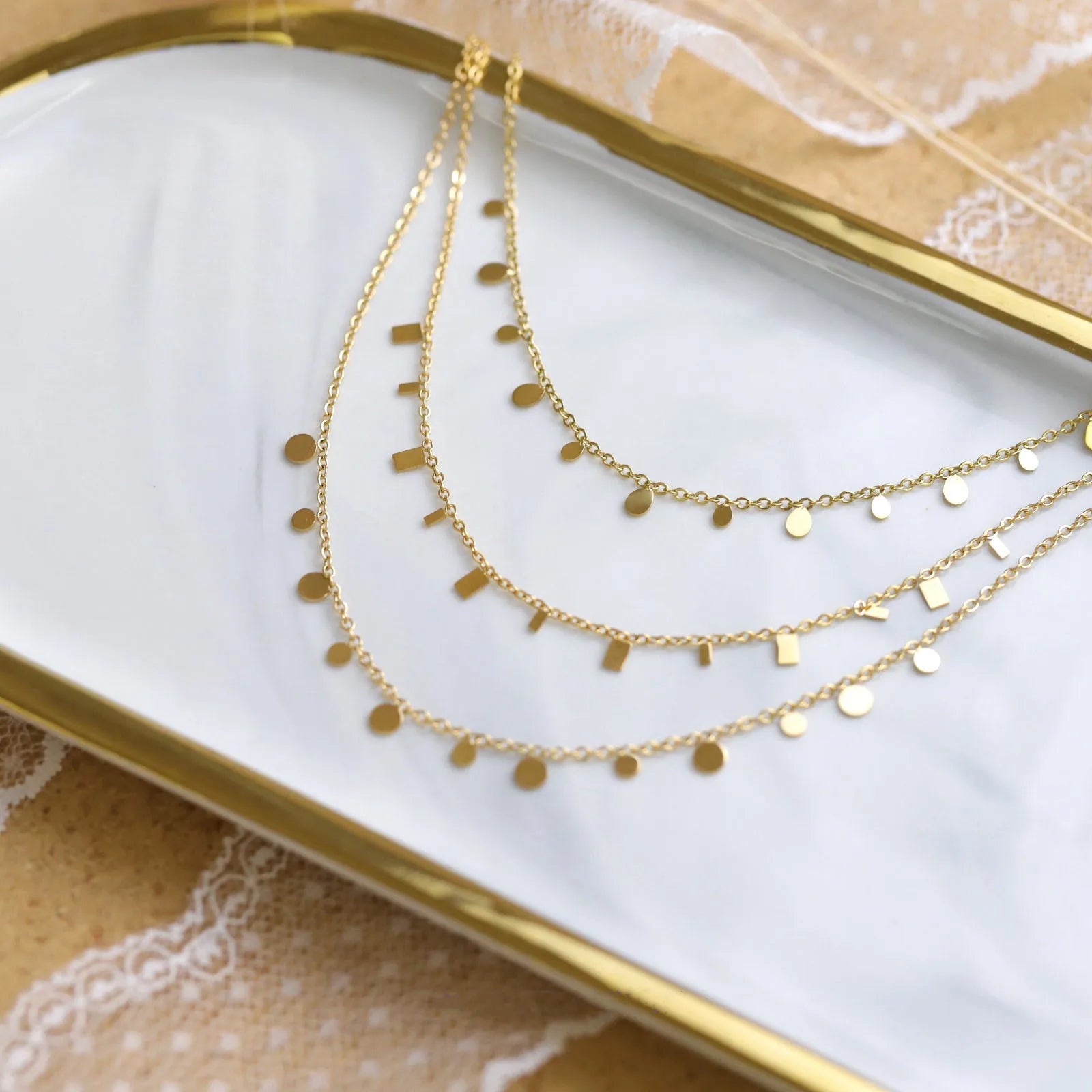 A three-layer necklace with delicate chains, featuring tiny alternating round disc and rectangular bar charms, draped elegantly on a white marble and tray. Dainty, minimalist, and modern fashion jewelry.