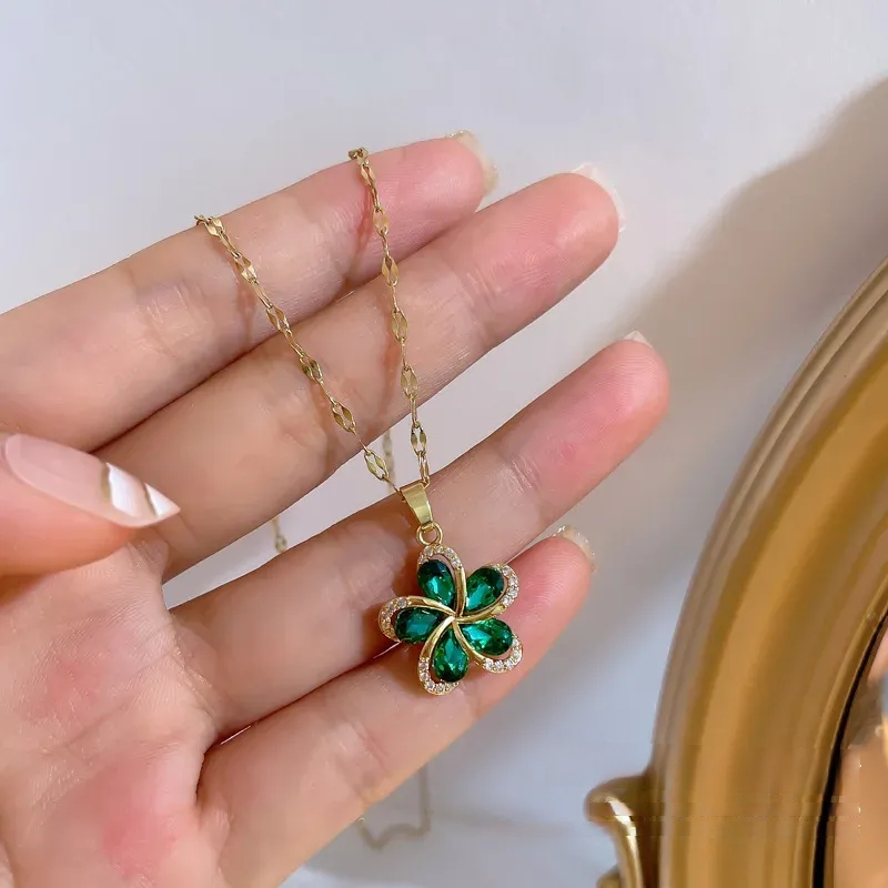 Close-up of a woman's hand holding a stylish gold necklace with a green flower pendant. The elegant floral design features emerald-like green stones and sparkling white accents, highlighting a beautiful piece of fashion jewelry