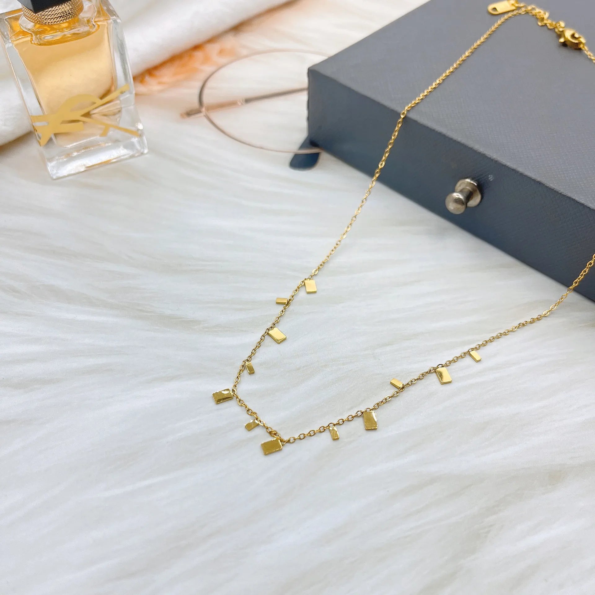 A delicate and minimalist golden necklace featuring small, rectangular charms is elegantly displayed on a soft, white, furry surface, accompanied by a perfume bottle, glasses, and a jewelry box, suggesting a stylish and thoughtful gift idea.