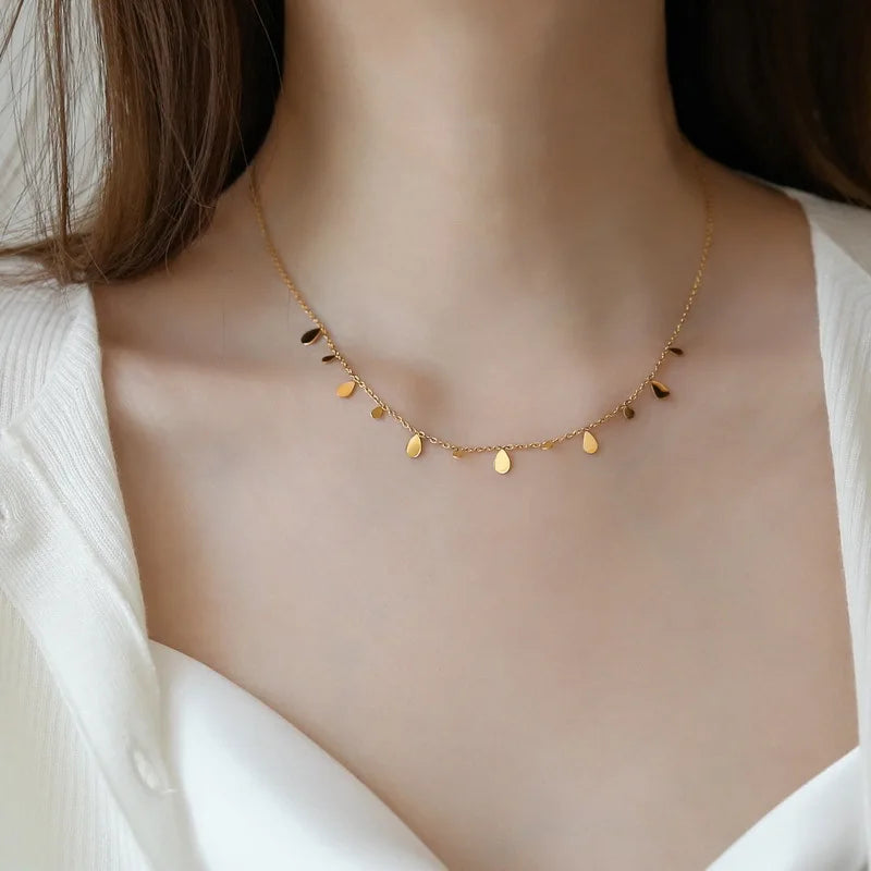 A delicate necklace with small, polished teardrop charms evenly spaced along the chain, worn by a woman with brown hair and a white top. Minimalist and elegant gold jewelry for everyday wear.