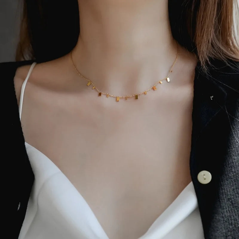 A delicate necklace with small, polished rectangular bar charms evenly spaced along the chain, worn by a woman with brown hair and a white top. Minimalist and modern gold jewelry for everyday wear.