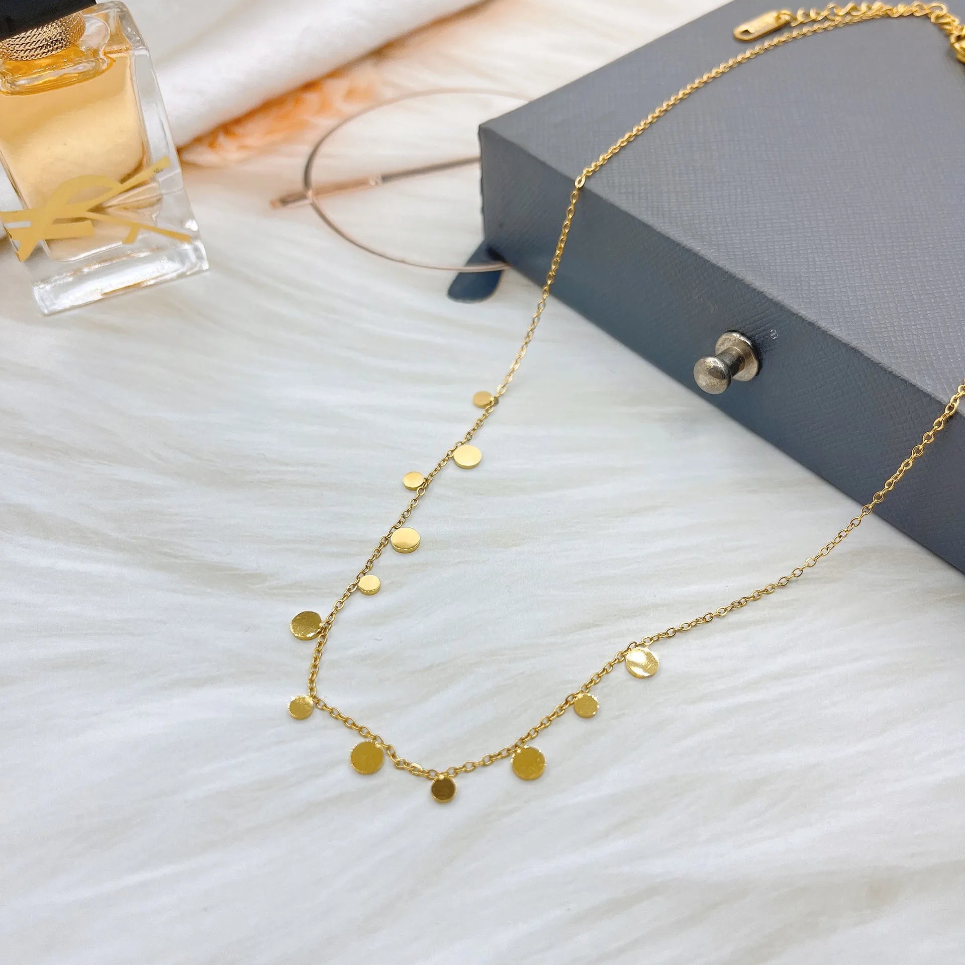 A delicate necklace with small, round coin charms is arranged on a soft white fur surface. In the background, a gray gift box, glasses, and a perfume bottle complete the stylish flat lay. Minimalist and elegant fashion jewelry.