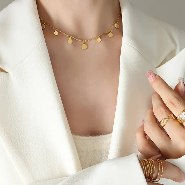 A delicate necklace with small, polished round coin charms, worn by a woman in a stylish white blazer. Her hands, adorned with several minimalist gold rings, are also visible. Elegant and modern jewelry for women.