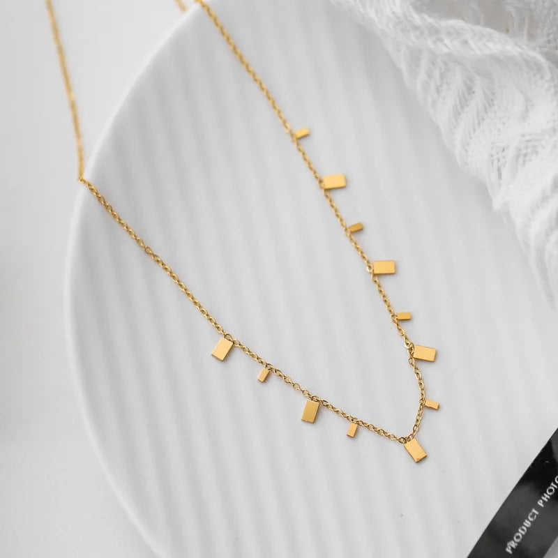 A delicate necklace featuring small, polished rectangular bar charms is artfully arranged on a simple white plate. This minimalist and modern jewelry piece is perfect for everyday style.