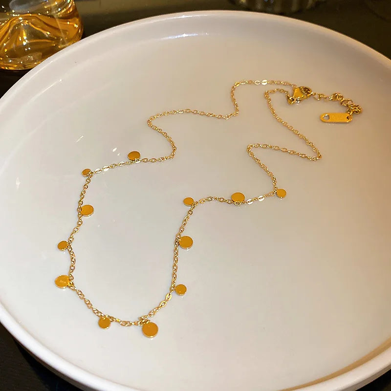 A delicate necklace featuring small, round coin or disc charms is artfully arranged on a simple white plate. This minimalist and modern jewelry piece is perfect for everyday style.