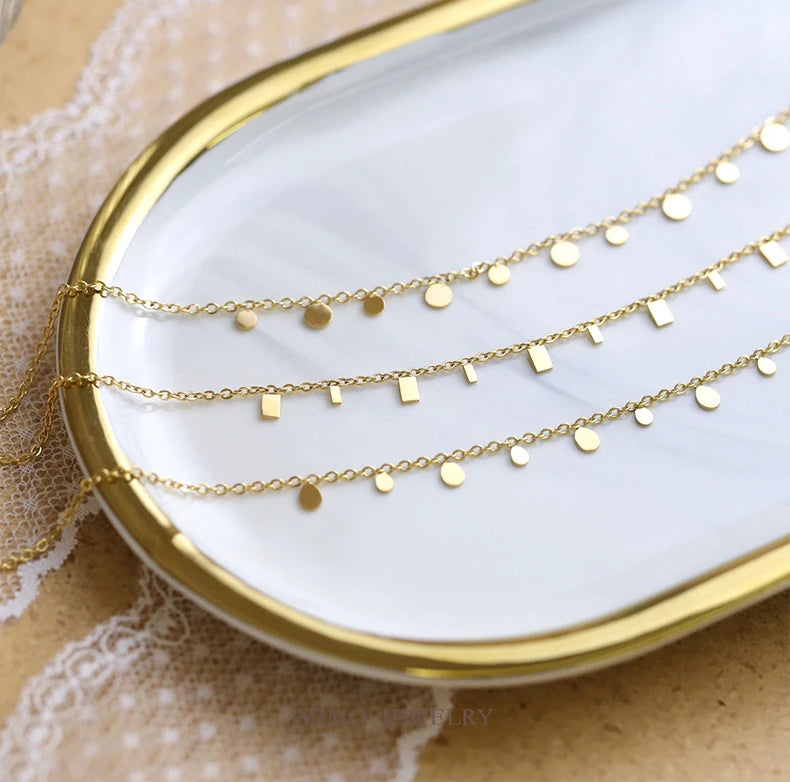 Athree-layer necklace with a combination of delicate chains. The top and bottom layers feature small round disc charms, while the middle layer has rectangular bar charms. The necklace is elegantly displayed on a white tray with a gold border, showcasing its modern, minimalist style.