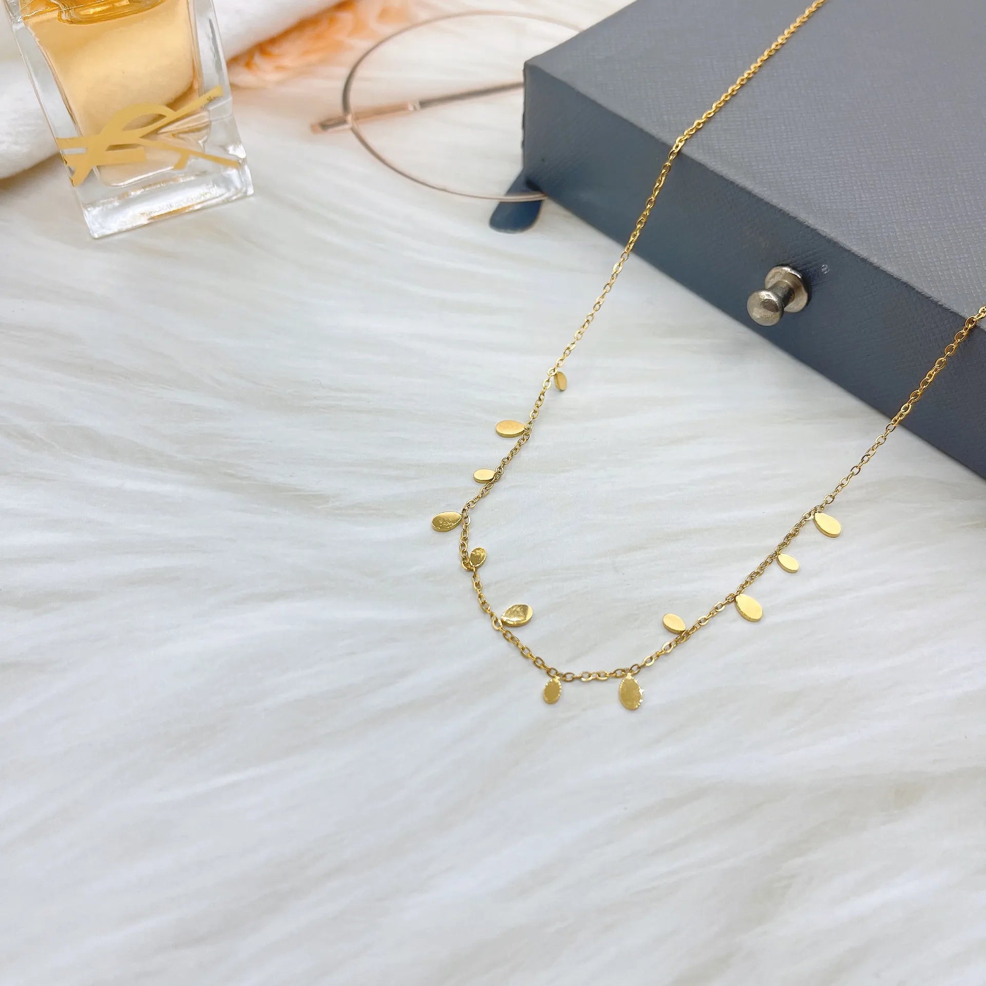 A delicate golden necklace with small, teardrop-shaped charms is arranged on a soft white fur surface. In the background, a gray gift box, glasses, and a perfume bottle complete the stylish flat lay. Minimalist and elegant fashion jewelry.