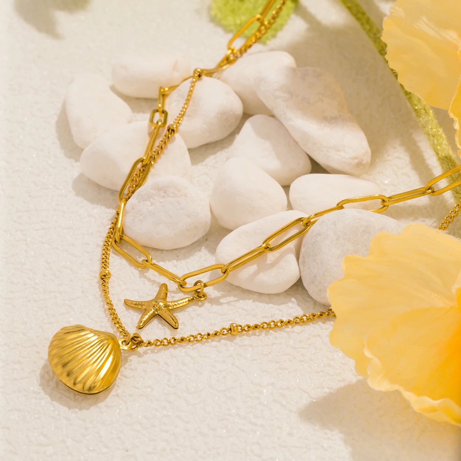 Detailed close-up of a stylish layered gold necklace featuring a delicate seashell and a charming starfish pendant, resting on smooth white pebbles with a soft yellow floral accent. Trendy summer jewelry and beach-themed accessories.