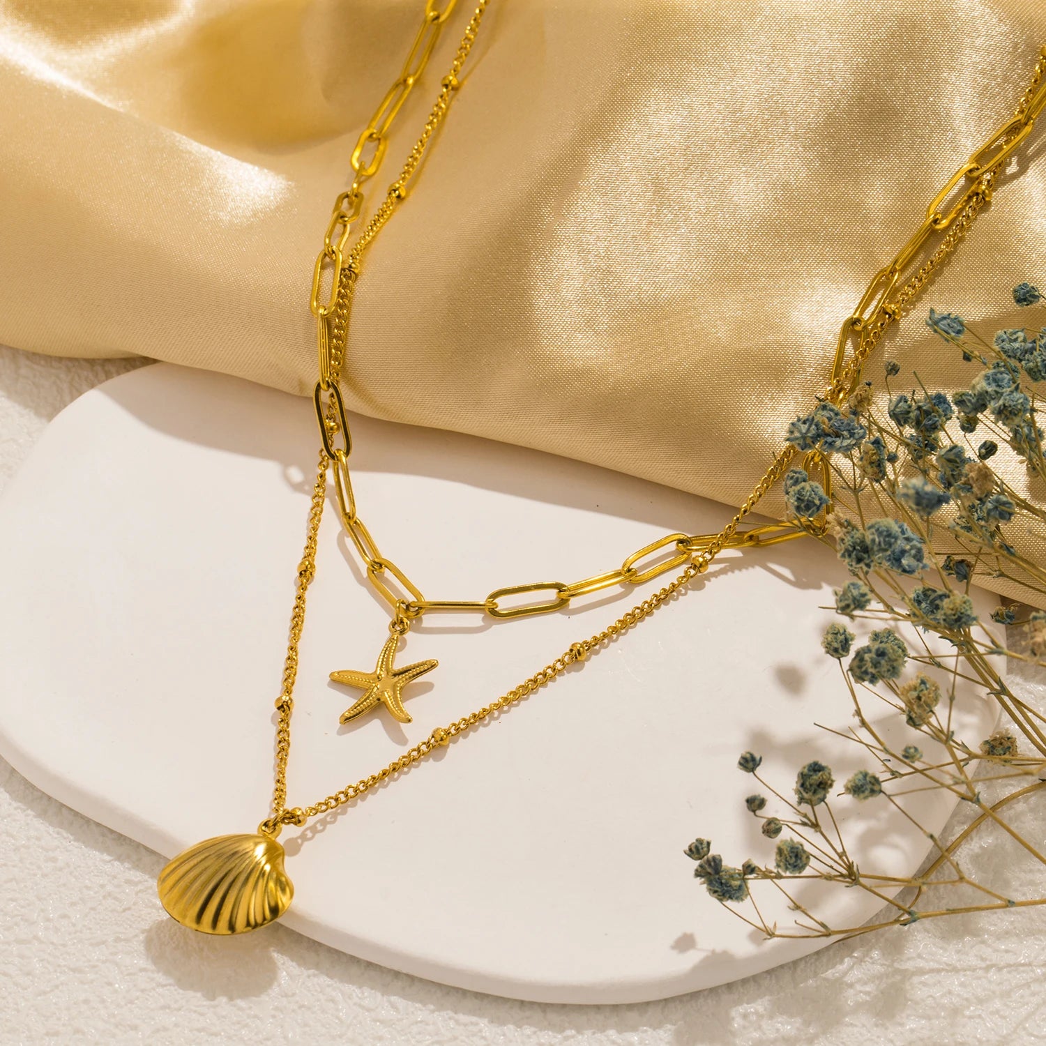 Elegant gold layered necklaces featuring a detailed seashell pendant and a small starfish charm, displayed against a soft, shimmering gold fabric and delicate blue flowers. Fashionable and dainty jewelry for women.