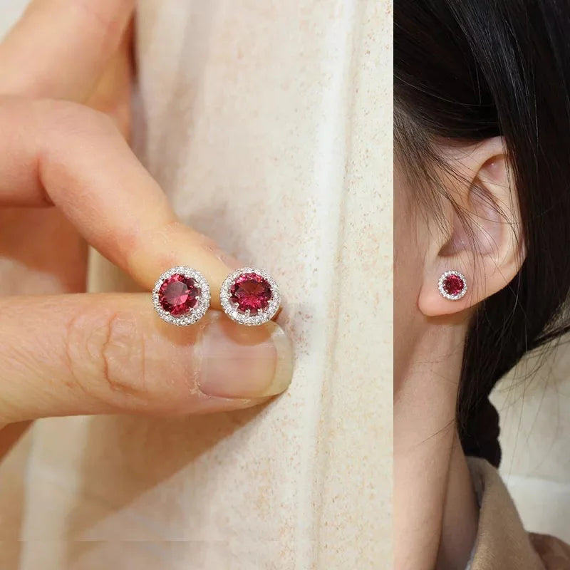 A pair of elegant round stud earrings featuring a rich red center gemstone encircled by a halo of sparkling clear stones is displayed in a split view, showcasing the earrings held by hand and worn on an ear, highlighting their vibrant color and sophisticated design.