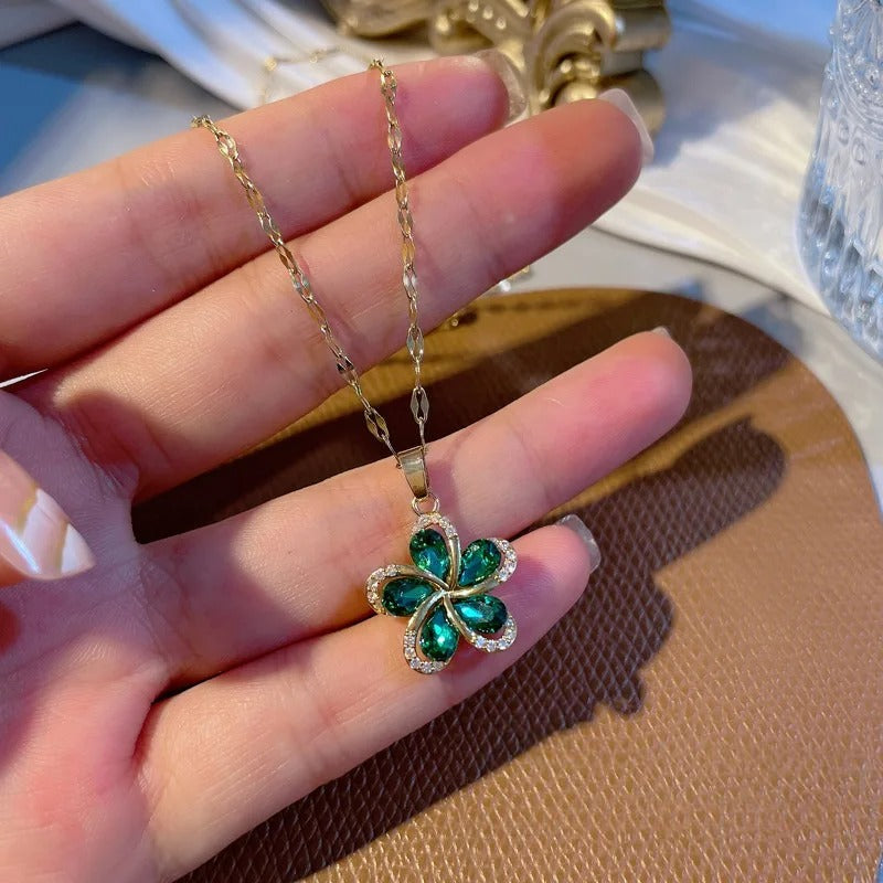 A woman's hand holding a delicate gold necklace with a green flower pendant featuring sparkling clear cubic zirconia stones. Close-up view of the elegant jewelry against a brown textured background.